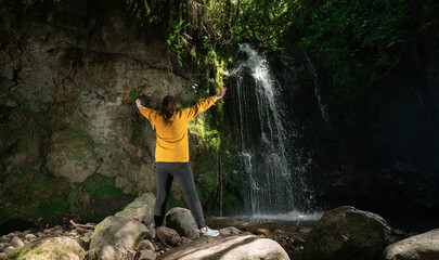 Woman dressed in a yellow jacket and black pants standing next to a waterfall, with her arms open, in the middle of a forest during a sunny day