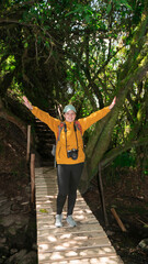 Woman dressed in a yellow jacket and black pants, carrying a red backpack, standing on a wooden bridge, with her arms open, in the middle of a forest during a sunny day