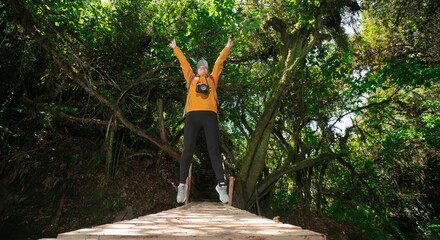 Woman dressed in a yellow jacket and black pants, jumping on a wooden bridge, with her arms open, in the middle of a forest during a sunny day