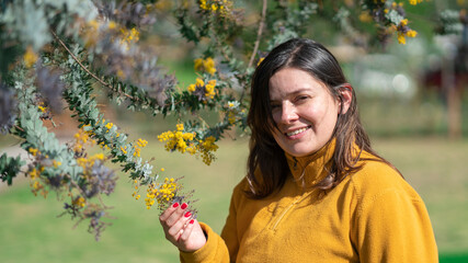 Woman smiling happily, dressed in a yellow jacket, standing next to a tree with small yellow flowers, during a sunny day