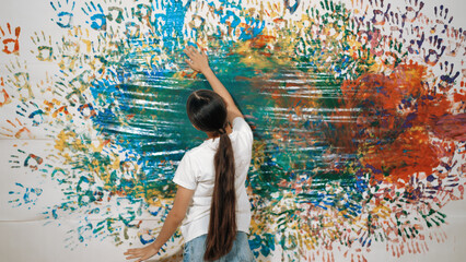Back view of young cute girl raise hand to paint the colorful stained wall. Attractive highschool student paint the wall with hand print while holding color palette. Creative activity. Edification. - Powered by Adobe