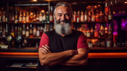Smiling bearded bartender standing with crossed arms in a well-stocked bar with a warm, inviting...