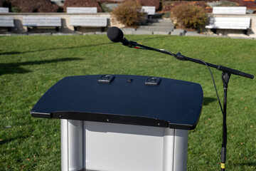 An outdoor park with vibrant green grass and a rock walkway. The area has been set up for a media scrum. A silver metal podium, black microphone, and two speakers on stands are ready for a speech.