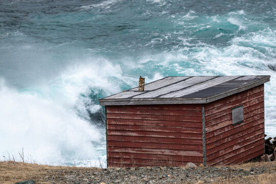 An old small red wooden shed with a stove pipe in the roof. There's a stormy sea in the background with teal teal ocean and white surf. The rough sea has a spray over the green rip curls of water.