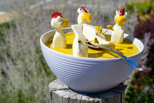 A colorful lawn ornament, chicken soup, made of white plastic. There are multiple small white and red-colored chickens with yellow beaks sitting in a yellow soup mixture with a spoon on top. 