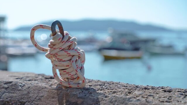 A person secures an anchor with a white and red coiled rope, with a scenic seascape and moored boats softly focused in the background