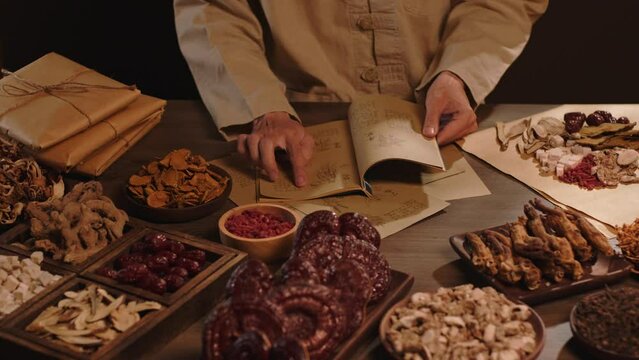 The physician is researching each type of medicinal herb in an ancient book, surrounded by wooden trays containing precious medicinal herbs. Scene from the front.