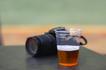 A cold beer in a clear plastic cup on the table, with the backdrop of a camera and lens, captures the essence of a relaxed evening.