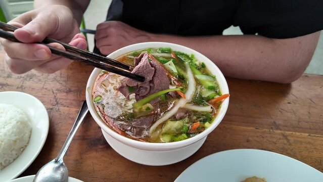 Person eating traditional Vietnamese pho soup with chopsticks, featuring sliced beef, noodles, and vegetables, with rice on the side