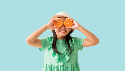 Happy Asian little girl posing with wear a hat with sunglasses holding orange slices on face, Holiday summer fashion green dress, isolated on pastel blue color background