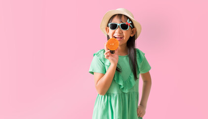 Happy Asian little girl posing with wear a hat with sunglasses holding orange slices, Holiday summer fashion green dress, isolated on pastel pink color background