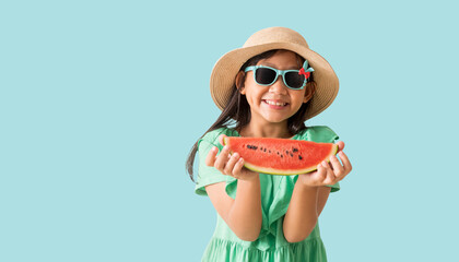 Happy Asian little girl posing with wear a hat with sunglasses holding watermelon slices, Holiday summer fashion green dress, isolated on pastel blue color background - obrazy, fototapety, plakaty