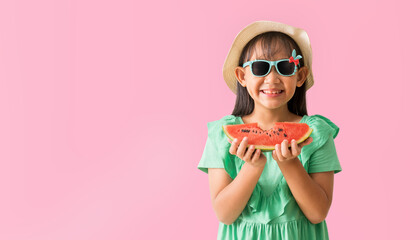 Happy Asian little girl posing with wear a hat with sunglasses holding watermelon slices, Holiday summer fashion green dress, isolated on pastel pink color background