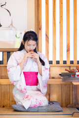 Pretty asian woman wearing Japanese traditional kimono.  Asian woman sitting near a low table.Young female enjoying time and drinking tea.Beautiful Woman smiling and sitting wooden chair..