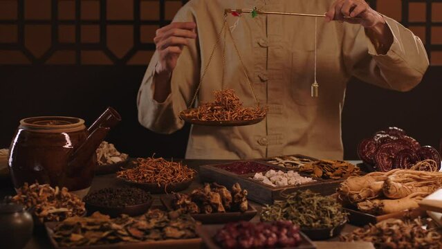 The physician is weighing dry medicinal herbs on a hand-held scale. Dry medicinal herbs are neatly stored in wooden trays, and a medicine pot is placed on the table.