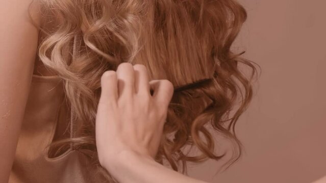 Woman hairdresser combing long wavy ginger female hair from back side.