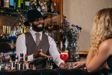 stylish black bartender offering a customer a cocktail decorated with garnish in a traditional cocktail bar