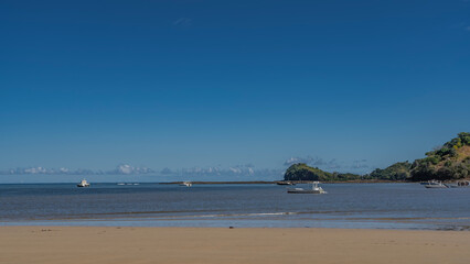 The boats are anchored in the calm blue ocean. Waves roll onto a sandy beach. A green hill against a clear azure sky. Light clouds over the horizon. Copy space. Madagascar. Nosy Be 