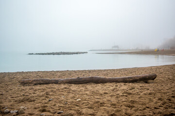 A foggy morning with beach sand and a horizontal log in foreground, water and a clam fog shrouded...