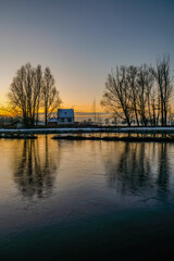 Fototapeta na wymiar This tranquil image encapsulates the serene beauty of a riverside cottage at dawn. The first light of the day casts a soft, golden glow on the scene, with the frozen river in the foreground reflecting