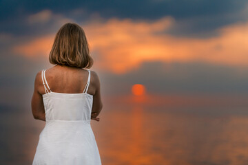 Fototapeta na wymiar A woman in a white dress stands on the beach, watching the sun set over the ocean