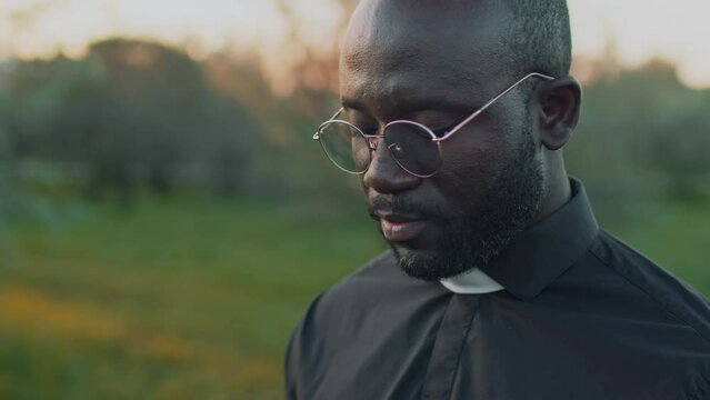 Tilt closeup of African American priest in glasses reading funeral service from prayer book at outdoor burial ceremony