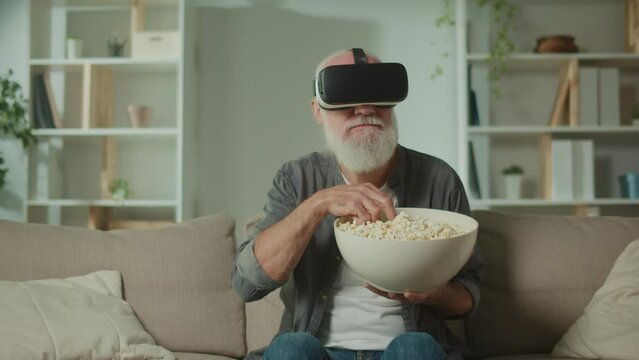 A Modern Old Man With Gray Beard in VR Glasses Examines Everything Around. An Elderly Man in VR Glasses Eats Popcorn. VR to Help the Elderly.