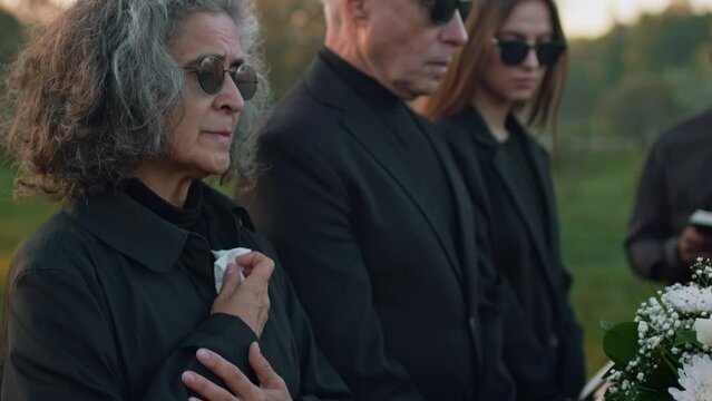 Tilt side footage of senior Caucasian woman in sunglasses holding back tears and using handkerchief standing with relatives by wooden coffin at outdoor funeral ceremony