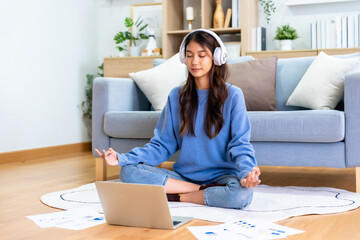 Happy young Asian woman practicing yoga and meditation at home sitting on floor in living room in lotus position and relaxing with closed eyes. Mindful meditation and wellbeing concept - obrazy, fototapety, plakaty