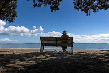Woman with hat sitting on a bench at the beach of Kohimarama beach Mission bay. Coast Auckland New Zealand