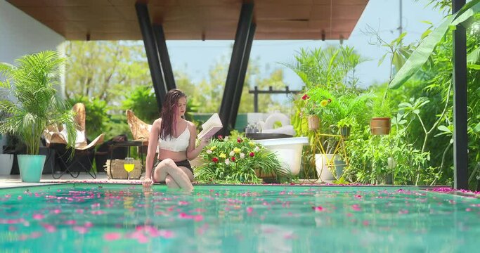 Indian Asian Hindu happy relax young lady woman sitting water pool edge read book pick up fresh drink hold juice glass outdoor home adult female girl enjoy fun joy luxury villa hotel resort trip day	