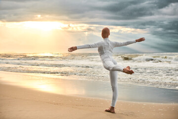 Young hairless ballerina with alopecia in white futuristic suit dancing on seashore at sunset sea, metaphoric surreal performance of bald pretty teenage girl exudes confidence, hope and unique beauty - obrazy, fototapety, plakaty