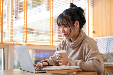 A positive Asian woman is sipping coffee while working on her laptop computer in a cafe.