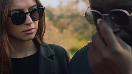 Side closeup of Caucasian young woman in sunglasses coming up to Black crying guy at cemetery and commiserating with him during funeral ceremony
