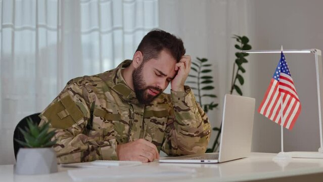 American bearded soldier feeling stressed and confused while working by computer next to American flag indoor. War veteran suffering from traumatic memories and trying to keep calm by massaging face.