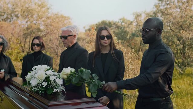 Tracking footage of Black guy coming to outdoor funeral ceremony, expressing condolences and putting white roses on coffin