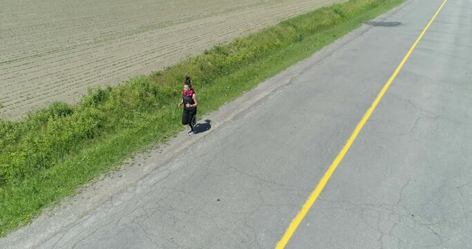 Drone flying backward in front of a woman jogging in a very long road in the country