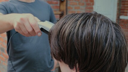the head of a person with long brown hair on a blurred background of a man in a gray T-shirt with a clipper in his hands in the courtyard of a private house in the process of cutting his hair at home