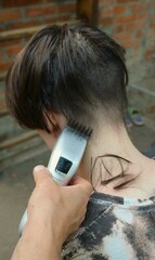 cutting a young man's hair with long dark hair using a clipper, back view, vertical image, a man's hand holds a trimmer with an attachment and cuts the back of a teenager's head