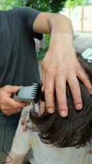 a man cuts the long hair of a teenager in the yard of a private house using a clipper, close-up, vertical image, family male relationships and household chores taking care of each other