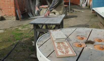 pieces of decorative tiles lying on a wooden surface on the street against a blurred background of a work process in which a craftsman processes tile material using a grinding machine