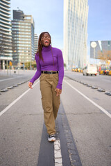 A joyful young woman in a purple turtleneck laughs while walking down an urban street