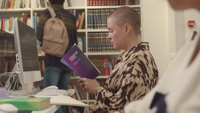 Medium shot of busy female Caucasian student with shaved head sitting in front of desktop computer with presentation on screen in college library and reading textbook on IT science