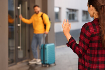 Long-distance relationship. Woman waving to her boyfriend with luggage near building outdoors