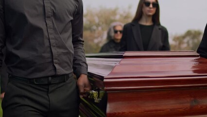 Midsection tracking footage of anonymous male and female pallbearers carrying wooden casket to resting place at cemetery during funeral ceremony