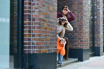 Three friends playfully spy on someone from around the corner of a building using binoculars. A comical portrayal of surveillance