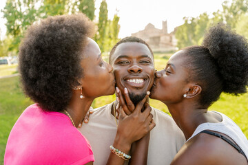 Two young beautiful african women with afro hairstyle kissing a happy smiling young man at the...