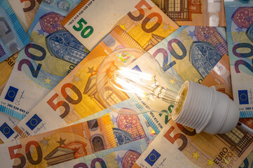 Light bulb lit, above euro banknotes. Electricity cost and tariffs.