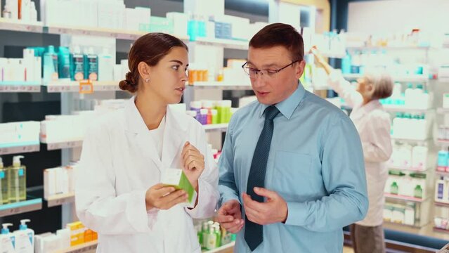 In pharmacy, girl apothecary hold package with cream and talks with man client about nuances of care. High quality 4k footage