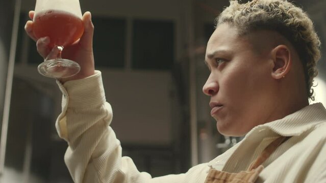 Tilt up o Biracial female brewmaster pouring glass of fresh lager from fermenter tank and taking sip while testing beverage produced at brewing factory